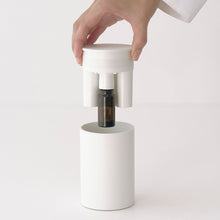 A hand in a white lab coat presses a white circular top onto a white cylindrical device, which releases a small amber glass bottle into the center. The scene is shot against a seamless, clean white background.