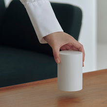 A hand in a white shirt lifting a plain white cylindrical container off a wooden table in a modern indoor setting.