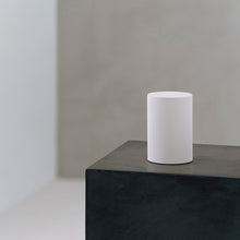 A minimalist white cylindrical table lamp sits on a dark textured pedestal against a neutral gray concrete wall background.