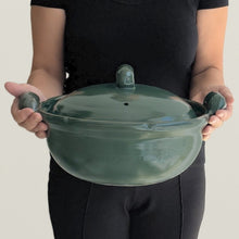 A person in a black outfit holds a dark green ceramic cooking pot with a lid and two small side handles against a plain light background.