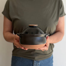A person in an olive green t-shirt holds a small black glazed ceramic pot with a lid and a natural terracotta base against a plain light background.